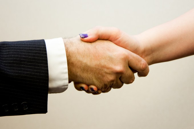 Shot of just hands as man and woman shake hands. He's wearing a black suit with white cuff showing. She has manicured finger nails with purple nail polish.