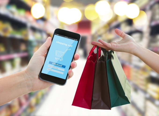 Man holding a smart phone with buy button while person hands him three small colorful shopping bags.