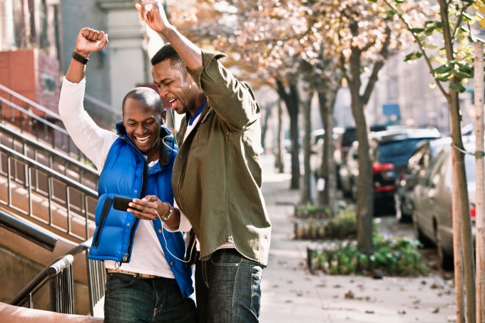 Two men using smart phone on street and gesturing with happiness