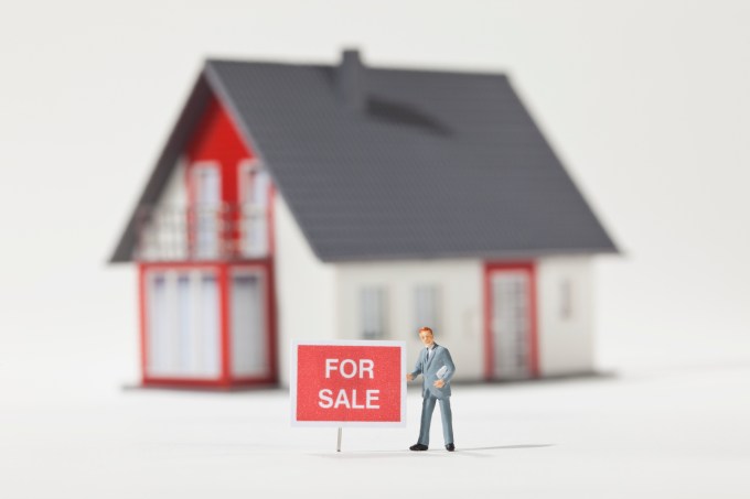 A miniature real estate agent figurine standing next to a FOR SALE sign